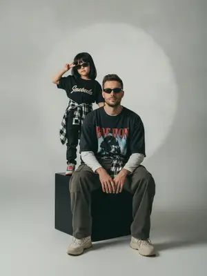 A photorealistic studio portrait captures a minimalistic editorial vibe, showcasing two individuals in a static scene. The model is seated on a large matte black cube, body slightly turned towards the camera, with knees bent and feet resting on the floor. Their arms are relaxed in front of their knees, fingers neatly extended. Standing behind and to the left on the same cube is a child, exuding confidence and playfulness. The child raises one hand in a "V" gesture while the other hand rests by their side, both figures sporting narrow dark sunglasses and directing their gaze towards the camera.

The composition features a frontal angle without noticeable perspective distortion, with the lens capturing a normal/short portrait view. The aperture is set around f/5.6–f/8, providing a moderate depth of field that keeps both subjects sharp while slightly softening the background. Soft studio lighting from a key source on the front left creates smooth transitions and gentle shadows against the backdrop, enhancing the overall aesthetic.

The background is a seamless light cyclorama with a prominent oval light spot in the center, surrounded by a subtle dark vignette. The studio setting is minimalistic, devoid of props, emphasizing a calm and stylish ambiance with a hint of retro street fashion and familial warmth. The seated model wears a dark oversized T-shirt with a bold print, layered with a light longsleeve and wide cargo trousers. They tie a plaid shirt around their waist and don chunky light sneakers. The child's attire mirrors the dark color scheme, with a T-shirt featuring a prominent chest inscription, paired with dark trousers, a plaid shirt, and colorful sneakers.

Details such as realistic fabric textures, natural creases, and fine folds add depth to the clothing, while the skin texture appears natural and unaltered. The scene is captured with high detail and sharpness, reminiscent of film photography with a light grain effect. The overall look is characterized by moderate contrast, a soft matte finish, and a focus on authenticity, free from overexposure or distracting elements. 
