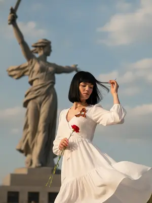 Фотосъемка возле монумента «Родина-мать зовет» в Волгограде. Камера смотрит вверх, немного наклонена. Женщина в кадре, вращается вправо. Волосы, ткань и пальцы с небольшим размытием от движения. Голова опущена, глаза закрыты, выражение спокойное. Рука у головы, расправляет прядь. Другая рука держит красный цветок. Белое платье с длинными рукавами, V-образным вырезом, рюшами и воздушной юбкой, развевается. На груди - Георгиевская лента. Вдалеке монумент «Родина-мать зовет» - женщина с мечом и поднятой рукой, голова повернута. Небо голубое, с облаками. Солнечный свет, теплые отблески, мягкие тени. Женщина справа внизу, монумент вверху. Цветокоррекция естественная, высокая детализация. 