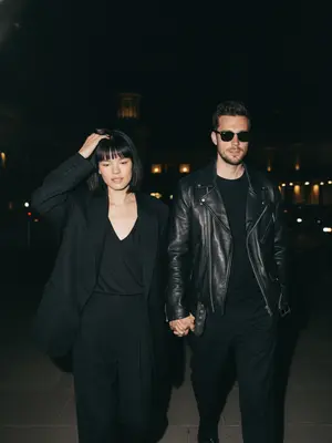 Capture a natural, paparazzi-style image under the night sky. A couple strolls together in the dimly lit street. The woman leads the way, casually adjusting her hair with a gentle gesture, while the man follows a bit behind, hand in hers. The camera captures the scene from a slightly elevated angle, zoomed in closely to the pair. Maintain the essence of a high-end celebrity magazine photograph. The facial features, eyes, nose, lips, hairline, and skin texture must stay true to their original form without any artificial alterations. 