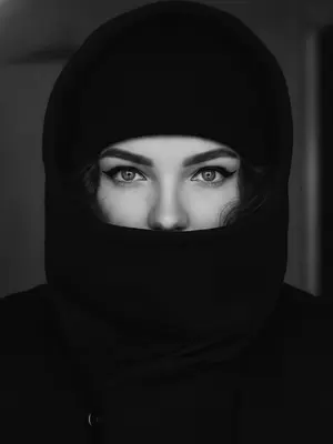 На переднем плане изображена женщина, ее лицо частично укрыто под капюшоном и темной шапкой-бини, которые создают загадочную атмосферу. Черная куртка с высоким воротником обрамляет нижнюю часть ее лица, открывая только яркие и выразительные глаза, которые притягивают внимание. Освещение мягкое, подчеркивающее черты ее лица и придающее сцене особую интимность. 