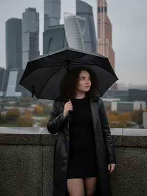 Городской пейзаж. На переднем плане изображена женщина, которая стоит у современного каменного перилла, с башнями «Москва-Сити» на заднем плане. Она смотрит вправо, опираясь на ограждение правым локтем, а корпус и ноги направлены к зрителю. В левой руке у нее раскрытый черный зонт, который защищает ее от дождя. Атмосфера сцены подчеркивается современными архитектурными линиями и динамикой городского окружения. 