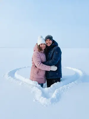 На фоне светло-голубого зимнего неба без облачка лежит плотный белоснежный снег, вырезанный в форме сердца. В середине этого сердца стоит пара, обнимаясь и улыбаясь в объектив камеры. Их стильная зимняя одежда выглядит современно, подчеркивая их хорошо видимые лица, на которых играют радостные улыбки.

Дневной свет придает снегу и тканям одежды мягкие холодные оттенки, подчеркивая каждую деталь. Атмосфера на фотографии наполнена уютом, любовью и спонтанностью зимнего момента. Снимок выполнен в реалистичном стиле, с фокусом на людях, создавая ощущение живого кадра, словно снятого с экшн-камеры или смартфона. Вертикальный формат добавляет динамики сцене.

Женщина одета в розовый пуховик с белой шапкой, украшенной помпоном, в то время как мужчина выбрал темный пуховик и шапку темного оттенка. 