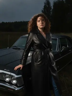 На фоне высокой травы, темных деревьев и серо-синего неба, модель в черном кожаном тренчкоте стоит возле винтажного автомобиля. Ее поза спокойна и уверенная, с легким разворотом корпуса к камере. Одна рука лежит на мокром капоте авто, вторая согнута у талии. Голова слегка наклонена, лицо выражает холодное спокойствие. Волосы растрепаны ветром, несколько прядей играют на лице. Тренчкот с широкими лацканами и поясом подчеркивает фигуру, кожа блестит от вспышки. Автомобиль показан с переднего угла, с каплей воды на капоте, фарами и стеклом. Камера расположена ниже лица, смотрит на модель с низу, кадрируя от бедра до головы. Вспышка создает четкие блики на лице, волосах и автомобиле, подчеркивая детали. Окружение остается темным, смешение света натурального сумеречного и вспышки. Фотография выполнена в стиле темного editorial fashion, с максимальным фотореализмом и глубокими тенями. 