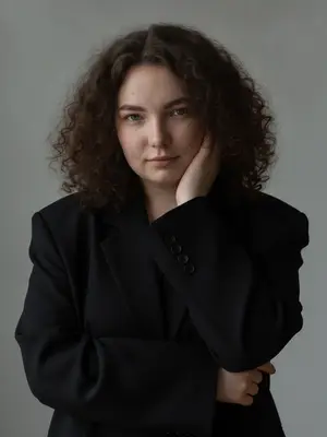 Крупный план, где в фокусе только лицо. Модель, напоминающая изображение, слегка наклонилась к объективу, ее плечи скрыты под широкими рукавами пиджака, создавая ощущение уюта. Обе руки находятся у лица: одна поддерживает щеку, а другая касается воротника, что придает позе интимность и защищенность. Голову она чуть опустила и повернута к камере, взгляд пронизывает объектив сквозь завитки волос, губы нежно сомкнуты, выражая легкую задумчивость и загадочность, с едва заметной улыбкой на лице. Атмосфера изображения наполнена таинственностью и глубиной. 