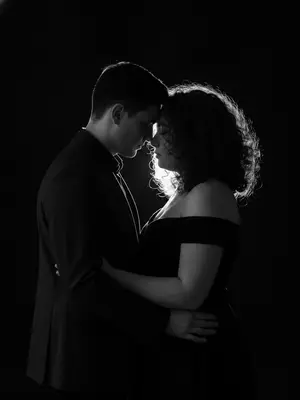 В темной студии создан фотореалистичный черно-белый портрет пары. Они стоят близко друг к другу, лбы и кончики носов практически касаются, словно в ожидании поцелуя. Мужчина, одетый в темную рубашку и пиджак, расположен слева, а женщина, в черном платье с открытыми плечами, стоит справа. Взгляды опущены: он устремлен вниз, к ее губам, а она либо закрывает глаза, либо направляет взор вниз. В их выражениях чувствуется нежность и спокойствие, без улыбок, лишь интимная атмосфера царит между ними.

Свет в студии создает жесткие контровые линии сзади и слегка сбоку, подчеркивая волосы и профили пары. Лица освещены мягкой полутенью, а в воздухе мелькают мельчайшие пылинки, освещенные светом. Фон полностью погружен во тьму, лишенный каких-либо деталей.

Оптика используется 85мм, с диафрагмой f/1.8-2.2, создавая малую глубину резкости с акцентом на линии носа и лба. Высокий контраст, глубокие черные тона и яркие световые пятна по волосам придают портрету кинематографичность. Пленочное зерно и легкое сияние по контуру добавляют изображению особую грань, сохраняя при этом натуральность и естественность черт лиц. 
