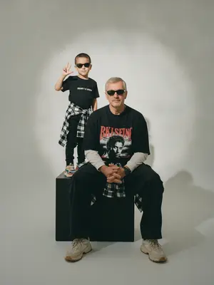 A photorealistic studio portrait captures a minimalistic editorial vibe, showcasing two individuals in a static scene. The model is seated on a large matte black cube, body slightly turned towards the camera, with knees bent and feet resting on the floor. Their arms are relaxed in front of their knees, fingers neatly extended. Standing behind and to the left on the same cube is a child, exuding confidence and playfulness. The child raises one hand in a "V" gesture while the other hand rests by their side, both figures sporting narrow dark sunglasses and directing their gaze towards the camera.

The composition features a frontal angle without noticeable perspective distortion, with the lens capturing a normal/short portrait view. The aperture is set around f/5.6–f/8, providing a moderate depth of field that keeps both subjects sharp while slightly softening the background. Soft studio lighting from a key source on the front left creates smooth transitions and gentle shadows against the backdrop, enhancing the overall aesthetic.

The background is a seamless light cyclorama with a prominent oval light spot in the center, surrounded by a subtle dark vignette. The studio setting is minimalistic, devoid of props, emphasizing a calm and stylish ambiance with a hint of retro street fashion and familial warmth. The seated model wears a dark oversized T-shirt with a bold print, layered with a light longsleeve and wide cargo trousers. They tie a plaid shirt around their waist and don chunky light sneakers. The child's attire mirrors the dark color scheme, with a T-shirt featuring a prominent chest inscription, paired with dark trousers, a plaid shirt, and colorful sneakers.

Details such as realistic fabric textures, natural creases, and fine folds add depth to the clothing, while the skin texture appears natural and unaltered. The scene is captured with high detail and sharpness, reminiscent of film photography with a light grain effect. The overall look is characterized by moderate contrast, a soft matte finish, and a focus on authenticity, free from overexposure or distracting elements. 
