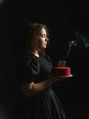 На изображении изображена женщина в фотореалистичном портрете с тем же лицом, чертами и выражением, что и на исходной фотографии. Ее цвет волос также идентичен. Сцена представляет собой день рождения, где мягкий свет свечи отражается на ее лице, а в воздухе плавают золотистые блестки, создавая атмосферу праздника. Фон окрашен в темно-серые тона. Освещение яркое и контрастное, с круглым световым пятном на заднем плане, бросающим выразительную тень. Волнистые волосы женщины мягко подсвечены. Она стоит немного повернутой в профиль и держит в руках небольшой праздничный торт, украшенный черными лентами и изысканной цифрой из золотых свечей. Женщина задувает свечу, излучая радость и веселье. 