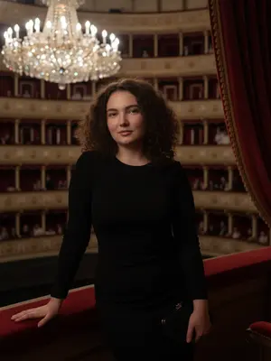 На фоне величественного Большого театра, я уютно расположилась в ложе, окруженной рядами других лож и огромной люстрой из хрусталя. Люди заполняют соседние места, а красные занавески из бархата создают атмосферу изысканности. Камера захватывает момент на уровне моих глаз, создавая впечатление, будто это фотография, сделанная на смартфоне: естественное освещение, нежная пикселизация, минимум ретуши, настоящий момент, неподдельный и живой. Мой взгляд направлен точно в объектив, с легкой улыбкой на губах. Голова слегка наклонена в сторону, придавая образу небрежную изящность. Волосы сверкают на свету, макияж природный, а губы блестят нежным глянцем. Одна рука ласково прикосается к краю ложа, а в другой у меня маленькая черная сумочка. Весь снимок окутан приглушенным светом, создающим атмосферу загадочности и элегантности. 