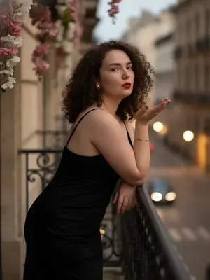 Стильный и реалистичный снимок, выполненный в уличной фотосессии с романтическим настроением. Женщина расположилась на узком балконе старинного здания, опираясь на темные металлические перила, которые имеют вертикальные прутья. На фоне виднеется размытая городская улица, освещенная вечерними огнями, а также светлая стена здания, украшенная свисающими розовыми и белыми цветами. Атмосфера кадра пронизана кинематографичностью, элегантностью и чувственностью, с легким налетом гламура. Цветовая палитра теплая и приглушенная, в которой преобладают темно-коричневые, бежевые и черные оттенки, а акцентом служит яркая красная помада на губах. Поза модели кокетливая и динамичная: она слегка наклонилась вперед, опираясь на перила, одна рука согнута в локте и поднята, словно она посылает воздушный поцелуй, а голова повернута вбок, лицо обращено к зрителю. На ее запястье сверкает тонкий золотой браслет, а в ушах блестят небольшие круглые серьги-пусеты. 