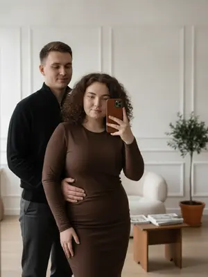 В уютной комнате с белыми стенами и изящной лепниной стоит мужчина и девушка. Она в обтягивающем коричневом платье с длинными рукавами, он в черном свитере с открытым замком на горле и темно-серых брюках. Мужчина обнимает ее за плечи и бедро, стоя за ее спиной. Девушка, стоя чуть впереди, держит в руке iPhone 17 в коричневом кожаном чехле, направив его на зеркало для селфи. 

Атмосфера в комнате светлая и уютная, с мягким освещением, создающим современный и элегантный вид. Цветовая гамма преимущественно состоит из нейтральных и теплых оттенков, где коричневый и черный играют главные роли. 

Позы и выражения лиц обоих говорят о дружелюбии, чувственности и спокойствии. Девушка и мужчина выглядят сближенными и расслабленными, взаимодействуя с легкостью. В заднем плане виднеется стильный мини-деревянный столик с журналами Kinfolk, мягкое светлое кресло букле и мини-оливковое дерево в горшке, создавая атмосферу уюта и изысканности. 