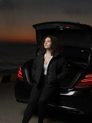 На краю морского пирса стоит черный Mercedes S-Class W222 AMG без номеров, блики луны играют на его кузове. В кадре виден только задний бампер и открытый багажник, из которого торчит часть модели. Девушка сидит на краю багажника, касаясь ладонью металлической детали, ноги покоятся на воздухе, обувь теряется в тени. Руки спрятаны в карманах кожаной куртки, голова слегка откинута назад. Ее взгляд устремлен вдаль, в сторону темно-оранжевого заката над горизонтом. Атмосфера пронизана тишиной позднего вечера, когда ночь уже на пороге. Освещение тусклое, с отсветом вспышки фотоаппарата, создающей реалистичные тени. Снимок сделан на Canon G7X, захватывая естественную позу в живом уличном кадре, словно картина ночного города. 