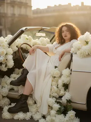A cinematic fashion editorial captures the essence of luxury and romance as a woman lounges gracefully inside a vintage convertible adorned with a lavish abundance of pure white flowers like roses, peonies, and hydrangeas, spilling over the seats and cascading along the edges.

The scene unfolds with the foreground dominated by the woman's legs, elegantly posed with one knee raised slightly higher, creating a sense of relaxed confidence. Her body leans back, head subtly tilted, and her gaze exudes a serene assurance, all while her arms rest naturally in a pose of effortless elegance.

Dressed in a light, flowing white or cream dress that billows softly around her, juxtaposed with chunky black leather combat boots, the styling combines elements of softness and edge. Her makeup features a bold red lipstick and thick lashes, enhancing her natural beauty.

Captured in the soft glow of the golden hour, the setting sun casts a warm, cinematic light on the scene, creating a dreamy atmosphere. The background showcases softly blurred architecture reminiscent of Parisian charm, adding a touch of sophistication to the composition.

The image is imbued with a romantic yet edgy mood, with a subtle interplay between softness and roughness. Every detail is meticulously rendered, from the realistic skin texture with visible pores to the intricate folds of the fabric and the delicate imperfections of the flower petals.

Shot with a 50mm lens and utilizing a shallow depth of field to create creamy bokeh, the photograph is ultra-detailed and photorealistic, capturing every nuance with precision. The lighting scheme combines the warm hues of the golden hour with a direct flash reminiscent of Y2K aesthetics, resulting in soft highlights and a slight haze that adds to the overall allure of the image. 