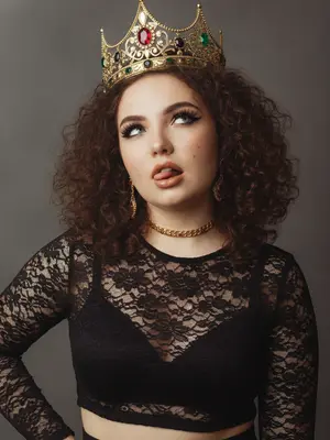 Описание портрета девушки, выполненного в смешанной эстетике «е-qirl» и «rebellious royalty»:

Девушка изображена в провокационной позе, выражающей дерзость и игривость. Ее глаза направлены в сторону и вверх, при этом кончик языка немного высунут, создавая образ с нотками бунта и отрешенности.

Макияж глаз выполнен с длинными острыми черными стрелками, густыми ресницами и хайлайтером, подчеркивающим внутренние уголки и скулы. Губы пухлые, окрашенные в телесный матовый оттенок с легким блеском и карандашом персиково-розового цвета.

На голове у девушки массивная корона в винтажном стиле с филигранным узором и драгоценными камнями: крупным рубином по центру и изумрудами и черными камнями по бокам. Корона сидит небрежно, добавляя образу неформальности.

Чокер на шее дополняет образ, сочетаясь с короной по стилю - цепочка с красными камнями. Серьги в ушах аналогично украшены.

Фон нейтральный, дымчато-серый, лишенный деталей, чтобы привлечь внимание к аксессуарам и лицу девушки. Освещение мягкое, рассеянное, с эффектом «зернистости», создающим винтажный вид. Цветовая гамма теплая, с акцентом на золото и красный.

Портрет транслирует современную интерпретацию власти и красоты через сочетание классических и современных элементов. Образ выглядит стильно, кинематографично и запоминающе, с короной золотого цвета. 