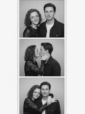 In a vintage black-and-white photobooth strip, a man and a woman are captured in four frames. The man dons a dark denim jacket over a light T-shirt, while the woman wears a fitted semi-sheer black long-sleeve top with delicate patterns. The energy between them is candid and authentic, with a natural, imperfect pose that feels unpolished and asymmetrical. The frames are stacked in a single column, each one showcasing a different moment. The background is a simple light studio setting, with white borders and square corners, evoking a realistic analog photobooth vibe.

In the first frame, they stand side by side facing the camera, the woman on the left and the man on the right. The woman's head is slightly turned to the right, with a flirtatious smile and a sidelong glance, creating a symmetrical yet slightly off-center composition. Moving to the second frame, the woman turns towards the man, gently cupping his cheek with a bright smile, while he looks back at her with a soft expression. Their closeness creates a left-weighted composition with empty space on the right.

In the third frame, the couple shares a kiss in profile, the woman on the left and the man on the right, positioned towards the left side of the frame with extra space on the right. Her hand rests on his cheek as they embrace. Finally, in the fourth frame, the woman hugs the man from behind over his shoulders, leaning her head towards him from the upper left corner, while he smiles at the camera. The couple is close to the left side of the frame, leaving the right half empty for a balanced yet intimate composition. 