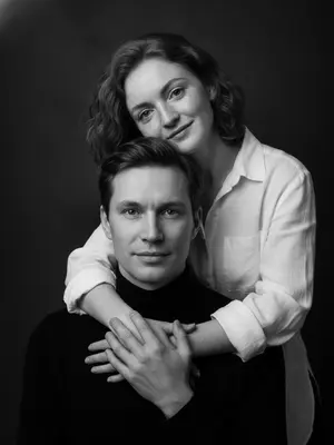 Изображение показывает родителя и ребенка, которые участвуют в семейной фотосессии. Фото выполнено в студии и имеет вертикальную композицию, с крупным планом лиц. Родитель, находящийся внизу кадра, направляет свой взгляд прямо в объектив, лицо полностью обращено к нам. Ребенок, находящийся сверху, повернул голову в сторону, его щека лежит на плече родителя, и он смотрит в камеру с нежной улыбкой. Рука ребенка обнимает шею родителя, видна в кадре, а родитель, в свою очередь, держит эту руку своей ладонью.

Одежда ребенка состоит из белой рубашки из льна, в то время как родитель одет в черную водолазку. Фон студии выполнен в темно-серых почти черных тонах, что создает контраст с яркими лицами на переднем плане. Освещение мягкое и боковое, оно придает изображению объем и глубину.

Фотография выполнена в черно-белой гамме с выразительными контрастами. Атмосфера, царящая на снимке, пропитана нежностью, интимностью и спокойствием. 