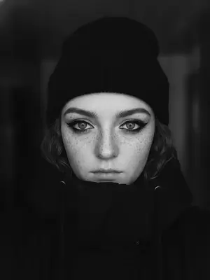На переднем плане изображена женщина, ее лицо частично укрыто под капюшоном и темной шапкой-бини, которые создают загадочную атмосферу. Черная куртка с высоким воротником обрамляет нижнюю часть ее лица, открывая только яркие и выразительные глаза, которые притягивают внимание. Освещение мягкое, подчеркивающее черты ее лица и придающее сцене особую интимность. 