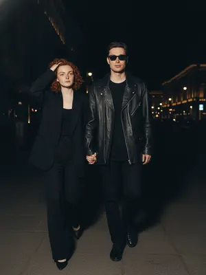 Capture a natural, paparazzi-style image under the night sky. A couple strolls together in the dimly lit street. The woman leads the way, casually adjusting her hair with a gentle gesture, while the man follows a bit behind, hand in hers. The camera captures the scene from a slightly elevated angle, zoomed in closely to the pair. Maintain the essence of a high-end celebrity magazine photograph. The facial features, eyes, nose, lips, hairline, and skin texture must stay true to their original form without any artificial alterations. 