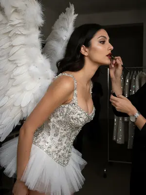 Создайте изысканную фотосессию в стиле Victoria's Secret. Молодая женщина стоит в профиль, слегка наклонившись вперед, готовясь к выходу на подиум. Визажисты наносят на нее помаду, их руки видны в кадре. На ней корсет, украшенный бисером и кристаллами, короткая пышная юбка и большие перьевые крылья. Изображение передает атмосферу «закулисья». Фон темно освещен, вероятно, это подиумное помещение. Основное внимание уделено лицу девушки и деталям ее наряда. Важно подчеркнуть выразительность взгляда и роскошь костюма. Фотография освещена вспышкой, что подчеркивает блеск бисера и кристаллов на корсете, а также сияющую кожу модели. Стиль Victoria's Secret: чувственность, роскошь, гламур. Разрешение 8K. Очень детализированное изображение. Важно сохранить исходное лицо модели. 