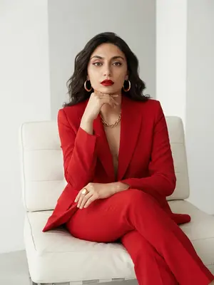 На фотографии, выполненной в стиле Vogue, запечатлены изысканная эстетика высокой моды и высокое разрешение. Женщина занимает позу на белом минималистичном кресле с прямыми широкими подлокотниками и низкой спинкой, обитой мягкой кожей. Фон яркий и минималистичный, подчеркивающий строгую геометрию сцены.

Ее поза выглядит расслабленной, но в то же время уверенной и собранной: корпус слегка повернут в сторону, ноги элегантно переплетены, одна рука покоится на колене с расслабленными пальцами, а другая согнута в локте, кисть приближается к лицу, а подбородок лежит на тыльной стороне ладони. Ее взгляд прямой, устремленный и уверенный, излучает силу и изысканность. Женщина повернута в профиль на три четверти, создавая эффект глубины и загадочности. 