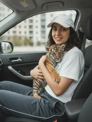 На фотографии должна быть изображена девушка, находящаяся в салоне автомобиля. Она одета в белую футболку, спортивные штаны и бейсболку. На снимке девушка держит в руках милого львенка. Улыбка на лице девушки должна быть естественной и приветливой. Важно сохранить реалистичность изображения и не изменять черты лица человека. 