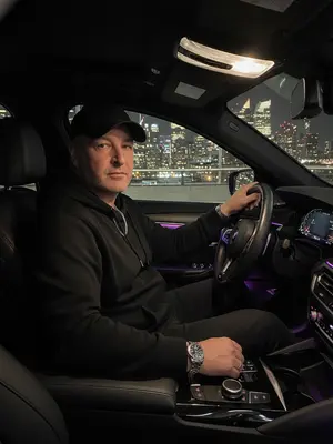Мужчина с элегантным образом за рулем роскошного BMW, стоит на фоне ночного города. Его рубашка тщательно заправлена, подчеркивая уверенность и стиль. Серебряные часы на запястье и цепочка на шее добавляют шик. Ладонью касается руля, взгляд направлен прямо в объектив с уверенностью. Салон автомобиля пропитан атмосферой роскоши: кожаные сиденья и яркие фиолетовые светодиоды создают уют. Вдали сверкают огни городской ночи, придающие образу стиль, статус и мощь. Съемка осуществлена на объективе 50mm f/1.8 с использованием вспышки, игра света и тени придают изображению выразительность. 