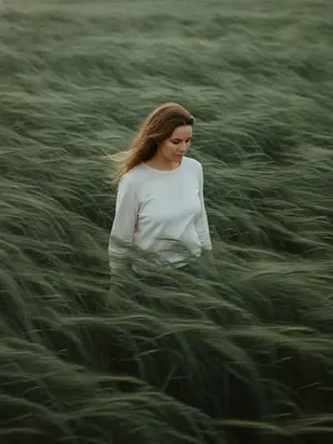 In a vast field of wind-swept sage green grass, a solitary figure stands amidst the flowing waves of tall grass. The subject wears a simple white shirt that contrasts softly with the lush green surroundings. Hair tousled by the wind, strands moving naturally in the breeze. With a gentle tilt of the head and eyes cast downward, a sense of quiet introspection and melancholy fills the atmosphere.

The expression is calm, distant, and restrained, capturing a mood of emotional depth. The frame is composed slightly off-center, from a subtle elevated angle, immersing the viewer in the texture of the grass without a visible horizon. The color palette is muted, with shades of sage green, deep olive, and soft off-white creating a natural and serene ambiance.

Shot from a telephoto perspective with a shallow depth of field, the cinematic framing enhances the editorial quality of the image. Film photography aesthetics add visible grain and a soft edge, while the grass sways with atmospheric motion blur driven by the wind. The scene tells a quiet, poetic story with ultra-detailed realism fit for a magazine cover. No text or watermark distracts from the pure visual narrative. 