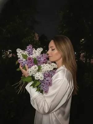 На темном фоне ночного города, тенистые деревья и майская листва создают задний план. Женщина стоит перед камерой, обе руки обнимают огромный букет сирени, белой и фиолетовой. Ее голова слегка наклонена к цветам, словно вдыхая их аромат. Макияж подчеркивает глаза черными стрелками и длинными ресницами, на губах блеск. Съемка происходит сбоку на уровне глаз, при слабом ночном освещении. Изображение высокой детализации, снятое на iPhone 17 Pro, передает живой момент с легким цифровым шумом и зернистостью, без использования вспышки. 