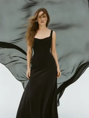 На студийном снимке fashion-портрета ультрареалистичного стиля, крупный план модели от груди до бедер. Модель стоит на белой циклораме, окруженной чистотой. За ней расправлена ткань из черного шифона, плавно колышущаяся под воздействием ветра. Этот элемент создает драматический фон с графичными контурами, ярко выделяясь на фоне белого пространства студии. 