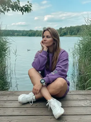 На деревянном пирсе у озера женщина стоит, обрамленная высокими камышами и зеленой листвой. В углу кадра ветка с листьями свисает. Озеро перед ней спокойно тянется, на фоне берега с деревьями под небом с белыми облаками. Утка плывет по воде вдали. Дневной свет придает цвета бирюзовые оттенки, делая кадр ярким и четким. 