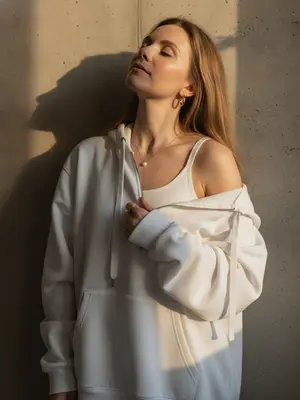 Женщина стоит у стены с текстурой, голова слегка наклонена назад, глаза закрыты, лицо повернуто к свету. Правая рука держит ткань толстовки у груди, а левая рука свободно опущена. Оверсайз толстовка с капюшоном спущена с правого плеча, видны завязки. На шее тонкая золотая цепочка с кулоном из жемчуга, золотые серьги среднего размера и кольцо на пальце. Макияж натуральный, с легким контуром, кожа сверкает естественным сиянием. Солнечный свет из окна создает резкие тени на стене и лице, добавляя контрастности. Съемка на объективе 50мм f/2.0, с малой глубиной резкости, цветовая палитра включает бежевый, золотой и серый. Фотография в стиле минимализма, с высоким фотореализмом и игрой теплых и холодных оттенков. 
