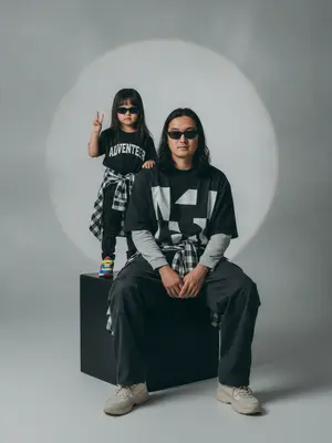 A photorealistic studio portrait captures a minimalistic editorial vibe, showcasing two individuals in a static scene. The model is seated on a large matte black cube, body slightly turned towards the camera, with knees bent and feet resting on the floor. Their arms are relaxed in front of their knees, fingers neatly extended. Standing behind and to the left on the same cube is a child, exuding confidence and playfulness. The child raises one hand in a "V" gesture while the other hand rests by their side, both figures sporting narrow dark sunglasses and directing their gaze towards the camera.

The composition features a frontal angle without noticeable perspective distortion, with the lens capturing a normal/short portrait view. The aperture is set around f/5.6–f/8, providing a moderate depth of field that keeps both subjects sharp while slightly softening the background. Soft studio lighting from a key source on the front left creates smooth transitions and gentle shadows against the backdrop, enhancing the overall aesthetic.

The background is a seamless light cyclorama with a prominent oval light spot in the center, surrounded by a subtle dark vignette. The studio setting is minimalistic, devoid of props, emphasizing a calm and stylish ambiance with a hint of retro street fashion and familial warmth. The seated model wears a dark oversized T-shirt with a bold print, layered with a light longsleeve and wide cargo trousers. They tie a plaid shirt around their waist and don chunky light sneakers. The child's attire mirrors the dark color scheme, with a T-shirt featuring a prominent chest inscription, paired with dark trousers, a plaid shirt, and colorful sneakers.

Details such as realistic fabric textures, natural creases, and fine folds add depth to the clothing, while the skin texture appears natural and unaltered. The scene is captured with high detail and sharpness, reminiscent of film photography with a light grain effect. The overall look is characterized by moderate contrast, a soft matte finish, and a focus on authenticity, free from overexposure or distracting elements. 