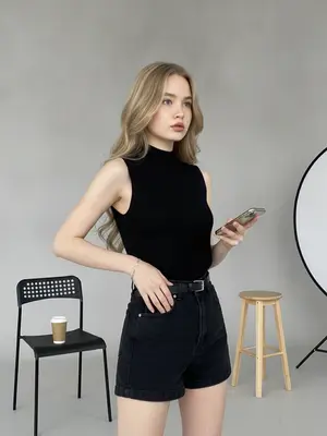 Фотосъемка проходит в светлой студии с минималистичным дизайном. На фоне видна серая стена и часть студийного оборудования, включая отражатель света и деревянный табурет. Цветовая палитра приглушенная, с преобладанием серых и нейтральных оттенков.

Модель стоит в полобороте, обращаясь вправо от объектива. Одна рука лежит на бедре, а другая держит мобильный телефон. На ней трикотажный топ без рукавов с высоким воротником и широкие брюки с высокой посадкой. Вокруг талии обвит тонкий черный кожаный пояс. На запястье блестит тонкий золотой браслет.

Рядом с моделью стоит черный стул, на котором стоит стаканчик с кофе. Обстановка создает атмосферу спокойствия и элегантности. 