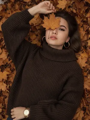 Фотореалистичное изображение, сделанное на iPhone. На плотном ковре из сухих кленовых листьев, окрашенных в золотистые, охристые, медные и коричневые тона, лежит женщина. Камера расположена чуть сбоку от ее тела, под углом, что позволяет увидеть ее голову в верхней части кадра, а корпус уходит вниз. Снимок близкий - женщина занимает почти все пространство, видна от макушки до середины груди, по краям остаются лишь немного листьев. Она расслаблена, голова слегка наклонена, лицо не строго анфас: подбородок повернут вбок, а взгляд направлен в объектив. Одна рука поднята, согнута в локте, и ее тонкие пальцы держат коричневый кленовый лист, который закрывает один глаз, в то время как второй глаз открытый и выразительный. На ней объемный темно-коричневый свитер оверсайз с крупной вязкой, свободно сидящий на плечах и корпусе, с высоким широким воротом и длинными рукавами. На поднятой руке, поверх свитера, находятся золотые часы с массивным металлическим браслетом, без дополнительных украшений. В ушах крупные эффектные круглые золотые серьги. Макияж включает длинные объемные ресницы с 2D-эффектом, а кожа имеет естественную текстуру с видимыми порами, без ретуши. Фон полностью состоит из осенних листьев, без лишних объектов, с немного пониженной экспозицией, натуральной цветопередачей и высокой детализацией, не размыт. 