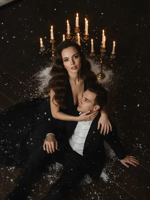 Съемка происходит с угла и сверху. В темной комнате разворачивается романтическая и драматичная сцена. На полу расположилась пара: мужчина и женщина, занявшая верхнее положение. Женщина, повернутая к объективу, поднимает лицо вверх, встречая взгляд камеры. Ее лицо просвечивает мягкий, теплый свет, выделяя выразительный, глубокий взгляд. Губы едва приоткрыты, и взгляд наполнен интригой. Волосы женщины, если они длинные, уложены в пышные волны, придающие образу голливудский шик. Если волосы короткие, они аккуратно уложены. На ней черное длинное платье с открытым лифом, подчеркивающее изящные линии и создающее драматический контраст. 