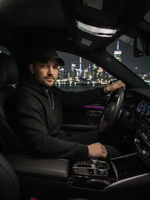 Мужчина с элегантным образом за рулем роскошного BMW, стоит на фоне ночного города. Его рубашка тщательно заправлена, подчеркивая уверенность и стиль. Серебряные часы на запястье и цепочка на шее добавляют шик. Ладонью касается руля, взгляд направлен прямо в объектив с уверенностью. Салон автомобиля пропитан атмосферой роскоши: кожаные сиденья и яркие фиолетовые светодиоды создают уют. Вдали сверкают огни городской ночи, придающие образу стиль, статус и мощь. Съемка осуществлена на объективе 50mm f/1.8 с использованием вспышки, игра света и тени придают изображению выразительность. 