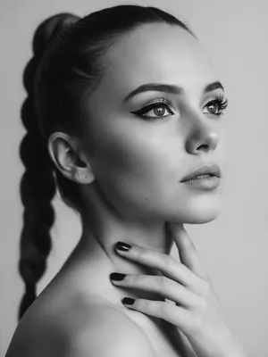Близкий крупный план в черно-белых тонах. Женщина изображена с плечами, лицо слегка повернуто в сторону и немного назад. Подбородок поднят, взгляд устремлен вверх и в сторону, не на камеру. Глаза выделены аккуратной длинной черной стрелкой, ресницы густые, создающие лисий эффект. Светлый оттенок и естественный контур подчеркивают скулы и форму лица. 