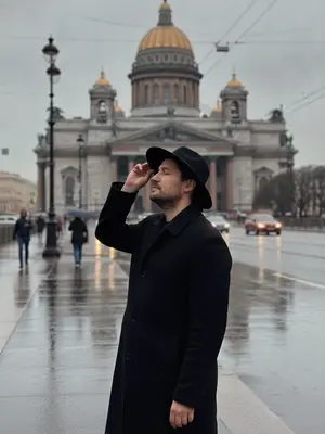 Фотография на улице Санкт-Петербурга после дождя, передающая атмосферу города. На снимке запечатлен человек в стильной зоне. Он стоит у фонарного столба, обрамленного сырым камнем, который отражает отблески влажного асфальта. Человек одет в черное пальто и широкополую шляпу, ладонью касается края головного убора. Его взгляд устремлен вдаль, словно он ожидает кого-то или что-то. Волосы чуть взмокли от оставшихся после дождя капель. На лице бледный свет от фонаря, создающий загадочную атмосферу. Вокруг витает запах свежести, а на фоне зданий виднеется туман, придающий картины загадочности и некой мистической притягательности. 