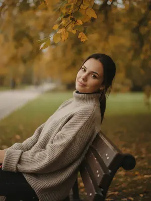 Осенняя фотосессия, запечатленная в вертикальном формате. На деревянной скамейке в парке расположилась женщина, удобно устроившись и немного откинувшись назад, ее голова слегка наклонена в сторону. Она смотрит прямо в объектив, на ее лице играет нежная улыбка. Уши украшены простыми кольцевыми серьгами. 