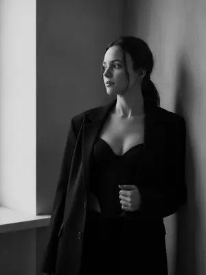 Женщина стоит в углу комнаты, наклонившись к стене. Ее лицо, с выражением спокойной уверенности, обращено к окну, вдалеке. На ней большой черный пиджак, небрежно надетый на плечи, и топ-корсет с V-образным вырезом. Рука лежит на пиджаке, поддерживая ее. Комната пронизана естественным светом из больших окон слева. Общая атмосфера внутри - стильная и спокойная, подчеркивающая элегантность и минимализм. Женщина позиционируется справа от центра кадра. Освещение мягкое, придающее ощущение простоты и минимализма. Черно-белая палитра фотографии выделяет контрасты и текстуры, создавая загадочную, но элегантную атмосферу. Портрет выполнен в ультрареалистичном 8K разрешении. 