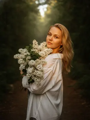 Фотография модели в лесу летом, снятая на Hasselblad X2D II 100C, 85mm объектив, f/1.8, крупный план, камера расположена на уровне талии и немного в стороне. Модель стоит на узкой тропинке среди густого леса, повернувшись через плечо к камере. В ее руках огромный букет цветов, который нежно обнимает, частично скрывая плечо и грудь. Лицо выражает спокойствие и чувственность, едва заметная улыбка, расслабленные губы, взгляд направлен прямо в объектив. На ней свободная белая длинная рубашка oversize с длинными рукавами, подсвеченная солнцем, ткань немного мятая. Кожа ровная, сверкающая, с легким загаром и текстурой, придающей реализма. Макияж мягкий и натуральный: легкое контурирование, пушистые брови, длинные ресницы, теплый свечение на коже. Теплый золотистый свет заката проникает сквозь листву, создавая кинематографичное освещение вокруг силуэта, играя на сирени и ткани одежды, образуя глубокие тени в лесу. Атмосфера тихая, живая и немного мечтательная, словно сцена из художественного фильма. Настроение задумчивое, с теплыми золотистыми оттенками, насыщенными зелеными тонами, мягким туманом, слегка зернистым эффектом, роскошным эстетическим видом в стиле Pinterest и фотореалистичным качеством RAW. 
