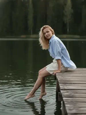 На фотографии, сделанной на iPhone, изображена модель, сидящая на деревянном пирсе у тихого лесного озера. Она присутствует в кадре в полный рост, фотография сделана со среднего расстояния. Камера расположена немного в стороне от пирса, на уровне талии модели, создавая непривычный ракурс. Пирс уходит в глубь справа, а вода и густой лес занимают основную часть фона, создавая атмосферу уединения и спокойствия. 