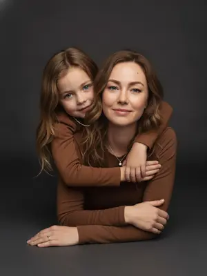 Create a studio portrait capturing the essence of a mother and child, maintaining their original facial features and expressions with precision. The scene is set in a close-up perspective at eye level. The duo is depicted lying on their stomachs, facing sideways to the camera. The child lovingly embraces the mother from behind, their arms wrapped tenderly around her shoulders. Both subjects gaze directly at the viewer, establishing a heartfelt connection. 

The mother exudes a natural beauty with a flawless, matte complexion complemented by subtle makeup - soft pink lips and a hint of mascara. She is dressed in a snug brown long-sleeved top, accentuated by a graceful silver chain and pendant draping elegantly along her collarbone. 

The child mirrors the mother's attire with their own brown long-sleeved top. They wear a delicate, thin chain necklace that harmonizes with the mother's jewelry, adding a touch of visual unity to the composition. 

The image is formatted in 9:16 ratio and rendered in stunning 8K quality. 