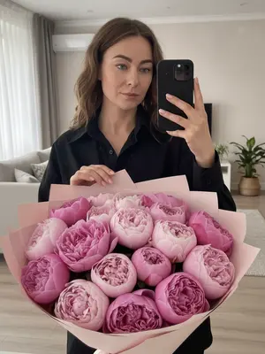 На снимке - отражение в зеркале, сделанное на смартфон iPhone 16 Pro. В центре кадра - девушка в черной рубашке с натуральным макияжем, длинными ресницами и блеском на губах. Ее правая рука держит телефон в черном чехле с камерой в углу, выделяющейся круглой формой объективов. Левой рукой она прикосается к поверхности за большим букетом, ногти аккуратно покрашены в светлый оттенок. На переднем плане виден огромный, пышный букет в пастельной упаковке, а на заднем плане - стильный домашний интерьер. 