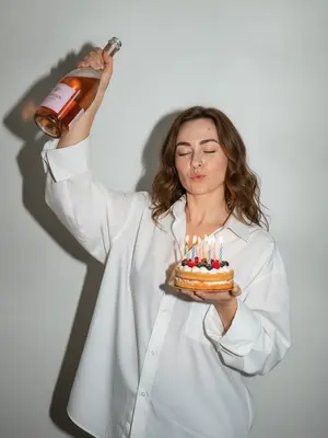 Изображение в стиле Party Girl / Birthday Aesthetic, сделанное в ультрареалистичной манере, как будто случайный снимок на старую цифровую камеру. Девушка в свободной белой рубашке оверсайз, застегнутой на пуговицы. В ее одной руке бутылка розового шампанского, которую она держит высоко, а другая рука держит небольшой бенто-торт с яркими свечами и свежими ягодами. 