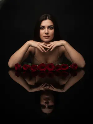 Женщина позирует в студийном пространстве, окруженная минимализмом. На блестящем черном столе отражаются ее силуэт и предметы. Фотография пропитана гламуром, подчеркивающим красоту. Вокруг царит спокойствие, с нотками таинственности. Цветовая палитра сочетает глубокий черный фон, ярко-красные розы и оттенки натуральной кожи. 