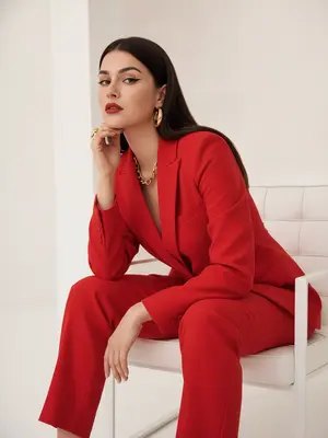 На фотографии, выполненной в стиле Vogue, запечатлены изысканная эстетика высокой моды и высокое разрешение. Женщина занимает позу на белом минималистичном кресле с прямыми широкими подлокотниками и низкой спинкой, обитой мягкой кожей. Фон яркий и минималистичный, подчеркивающий строгую геометрию сцены.

Ее поза выглядит расслабленной, но в то же время уверенной и собранной: корпус слегка повернут в сторону, ноги элегантно переплетены, одна рука покоится на колене с расслабленными пальцами, а другая согнута в локте, кисть приближается к лицу, а подбородок лежит на тыльной стороне ладони. Ее взгляд прямой, устремленный и уверенный, излучает силу и изысканность. Женщина повернута в профиль на три четверти, создавая эффект глубины и загадочности. 