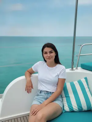На палубе катамарана модель сидит удобно, одна рука отдыхает на белом борту, а другая лежит на ноге. Ее корпус немного повернут к камере, глаза смотрят прямо в объектив, лицо украшает спокойная улыбка. Она одета в белую футболку и шорты.

Камера находится на уровне груди или немного ниже, а в кадре видно большую часть тела модели и окружающей среды. Фон занимает большую часть кадра, с небом в верхней части. Горизонтальная линия делит кадр пополам. Справа от модели виден кусочек катамарана с металлическими опорами и бирюзовым сиденьем, а на палубе заметна подушка с бирюзово-белыми полосами.

Используется стандартный объектив с фокусным расстоянием около 35–50 мм и диафрагмой примерно f/4–f/5.6. Глубина резкости умеренная: модель четко в фокусе, а фон (море и небо) слегка размыт, без явного боке. 