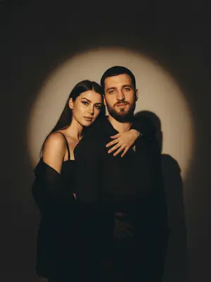 В студийном свете создан фотореалистичный портрет пары, олицетворяющий драматичную Y2K fashion-эстетику. Мужчина стоит на переднем плане, его корпус легко повернут в сторону, выставляя одно плечо ближе к камере. Поза его наполнена спокойствием и уверенностью, в то время как женщина стоит немного ниже, плавно обнимая его за шею и плечо. Ее лицо, выражающее дерзость и интимность, видно из-за его плеча, создавая эффект editorial стиля.

Женщина выглядит выразительно и гламурно, с cat-eye макияжем, четко очерченными губами и хайлайтером на коже. Она одета в черный корсет и оверсайз-пиджак с открытым плечом, в то время как мужчина носит черный лонгслив. Фон студии окрашен в темные оттенки, практически черный, с мощным прожектором, создающим большой круг света за спинами персонажей.

Свет от прожектора мягко освещает фон, растворяясь по краям и погружаясь в темноту, а персонажи частично перекрывают его, проецируя жесткие тени на стену. Освещение фронтальное, с высоким контрастом, реалистичными бликами и глубокими тенями на коже, а также легким пленочным зерном. Камера уровня глаз, с портретным объективом и плотным кадрированием, захватывает каждую деталь в 8K RAW качестве.

На снимке отсутствуют лишние элементы, текст и логотипы, подчеркивая исключительно модные образы и драматичную атмосферу. 