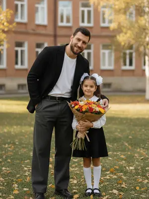 На улице запечатлен трогательный момент между родителем и ребенком, стоящими перед школьным зданием. Родитель, слегка наклонившись в сторону малыша, обнимает его одной рукой, прижимая к себе. Оба с искренними улыбками смотрят в объектив, создавая атмосферу тепла и близости. Их позы выглядят естественно и непринужденно, словно это не фотосессия, а мгновение из жизни. 