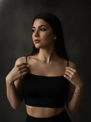 Фотография в стиле moody boudoir editorial, воплощающая атмосферу интимности, чувственности и приглушенного драматизма, представляет собой крупный план по пояс. Женщина стоит в полумраке, ладонями касается края черной ткани, изящно придерживая ее. Ее плечи слегка приподняты, а руки с длинным светлым маникюром сжимают тонкие бретели топа. Голова немного повернута в сторону, взгляд задумчиво устремлен вдаль, а выражение лица томное, загадочное и уверенное.

Вечерний макияж добавляет выразительности: гламурные стрелки, густые ресницы, создающие эффект «кошачьего взгляда», мягкий контуринг лица и матовая помада цвета пыльной розы. Женщина одета в облегающий черный кроп-топ на тонких бретелях-спагетти из матовой эластичной ткани, дополненный изящным серебряным браслетом на запястье.

Интерьер минималистичен, в темных тонах, с однотонным фоном, слегка текстурированным, погруженным в глубокую тень, создающую камерную обстановку. Низкоключевое освещение мягко падает сбоку, подчеркивая рельеф ключиц, линию плеч и текстуру кожи, создавая объем и драматичное настроение.

Используя объектив 85мм f/1.4, фотограф добивается идеальной фокусировки на глазах и лице модели, смазывая плечи и руки в мягком размытии. Гиперреалистичная текстура кожи, видимые поры, естественный блеск и отсутствие пластичности после ретуши создают естественный образ.

Общая эстетика фотографии - "dark feminine energy", создает интимную, теплую и дорогую атмосферу, наполненную мистикой и загадкой. 